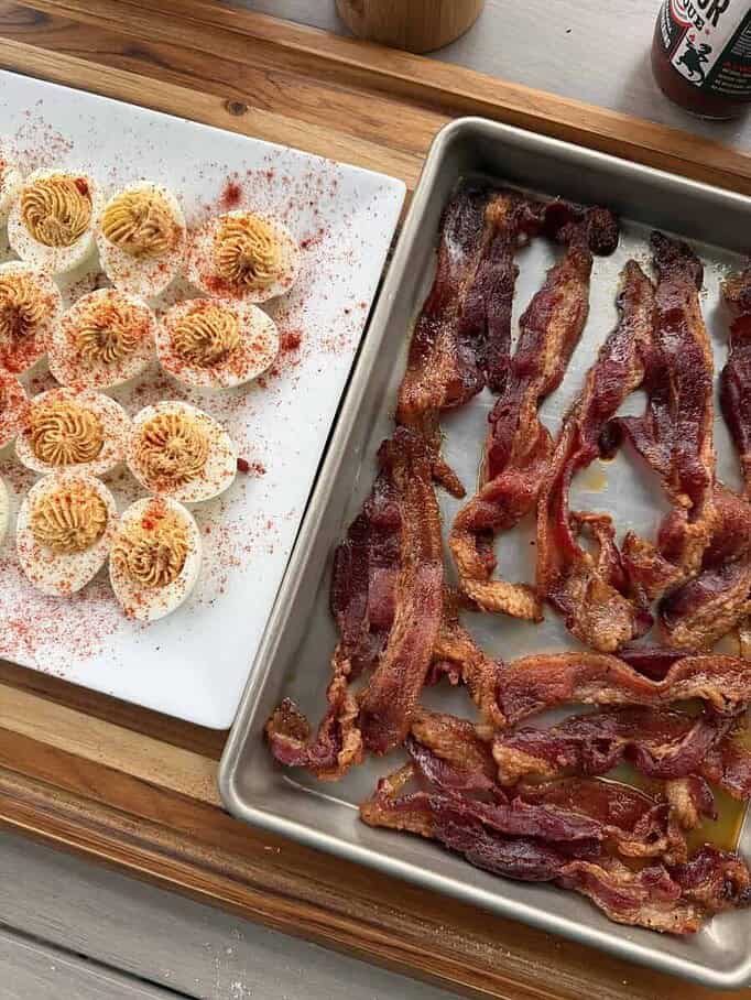 A plate of deviled eggs with sprinkled paprika on top alongside a tray of cooked candied bacon